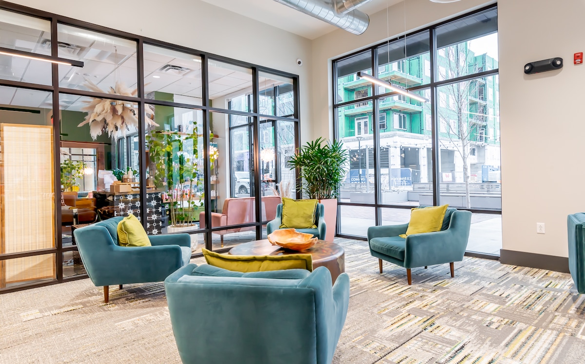 A modern shared living space is depicted, featuring soft teal armchairs arranged around a wooden coffee table. Lush greenery can be seen in the background alongside large windows, providing ample natural light. The interior design reflects a welcoming, relaxed atmosphere.