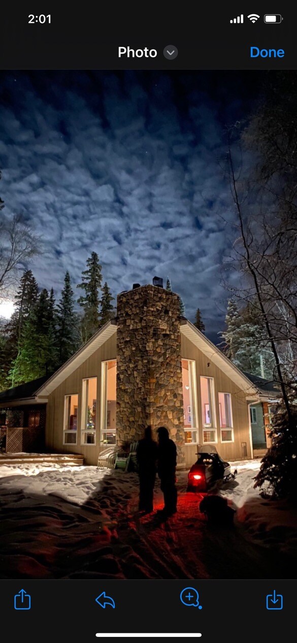 The exterior of a cozy lakefront home is framed by tall trees under a cloudy night sky. A prominent stone chimney adds character to the structure, while a gentle snow blanket covers the ground. Two figures stand in the foreground, silhouetted against the warm glow of illuminated windows.