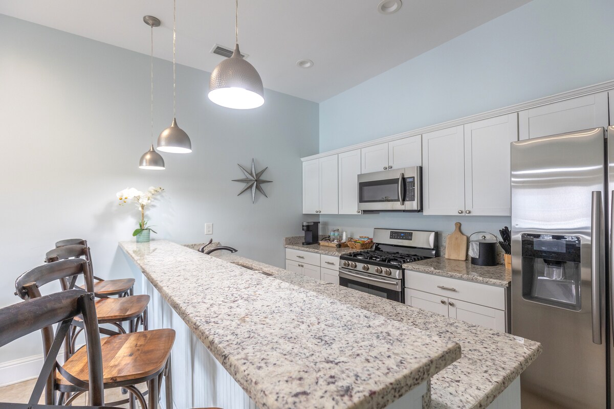 A modern kitchen features a long granite countertop with bar stools positioned alongside. Sleek cabinetry and stainless steel appliances are visible. Pendant lights hang above, illuminating the space, while a decorative star accent is mounted on the wall.
