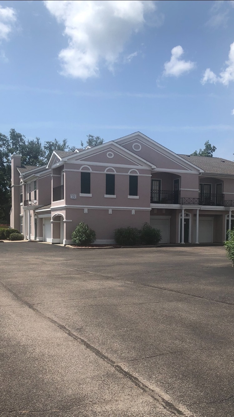 The exterior of a multi-story building is presented, featuring a light pink façade with decorative white accents. Large windows allow natural light to enter, and a balcony is visible on the upper level. Lush greenery surrounds the base of the structure.