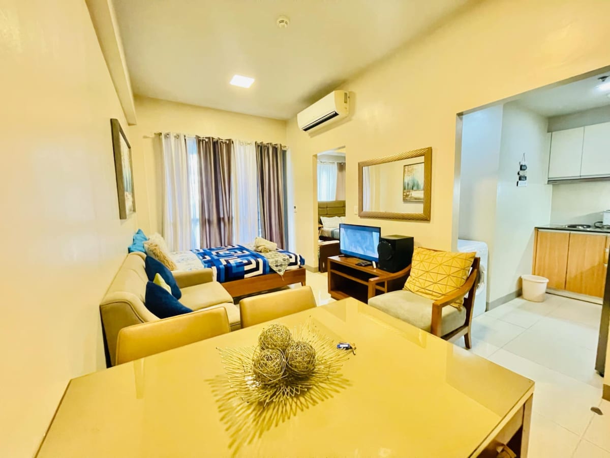 A bright living area is visible, featuring a cozy sofa and a dining table. A flat-screen TV sits across from the sofa, while a queen-sized bed is arranged with linens in the background. Natural light enters through the large windows adorned with light curtains.