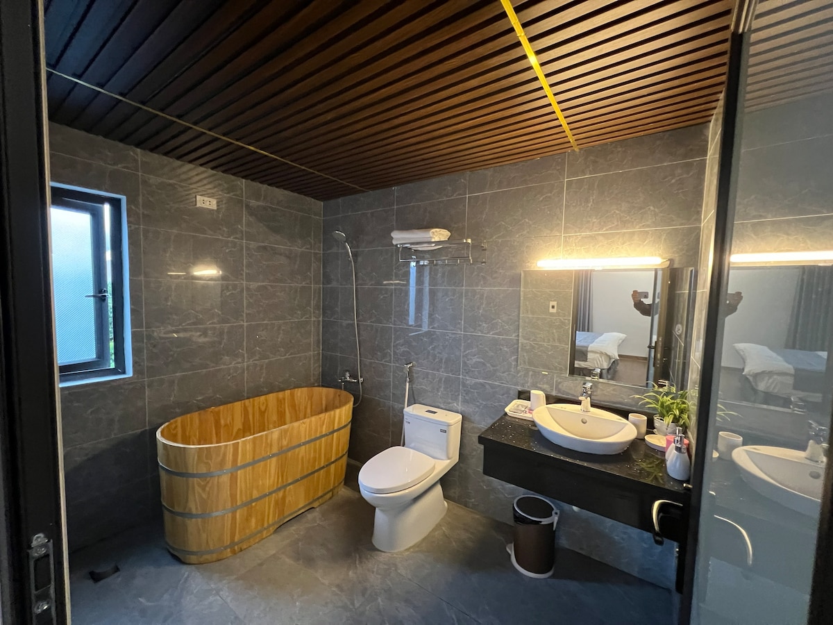 A modern bathroom features a wooden soaking tub alongside a sleek toilet and a contemporary sink with a round mirror above. Natural light enters through a small window, while a dark stone tile floor complements the overall minimalist design.