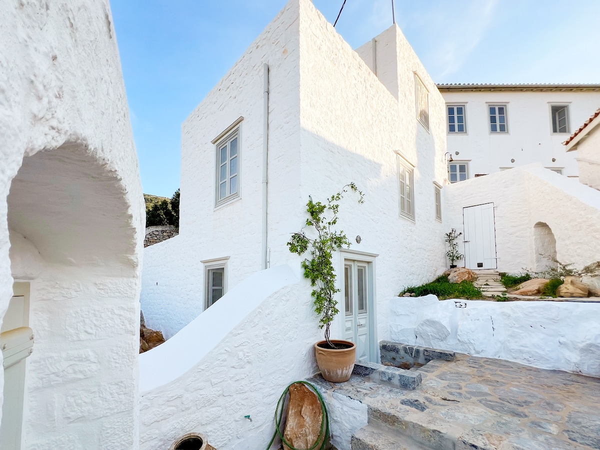 A charming white stone house is situated at the edge of a hillside, featuring multiple levels and large windows. Plants are seen in terracotta pots near the entrance, while stone steps lead to the doorway, blending the home with its tranquil natural surroundings.