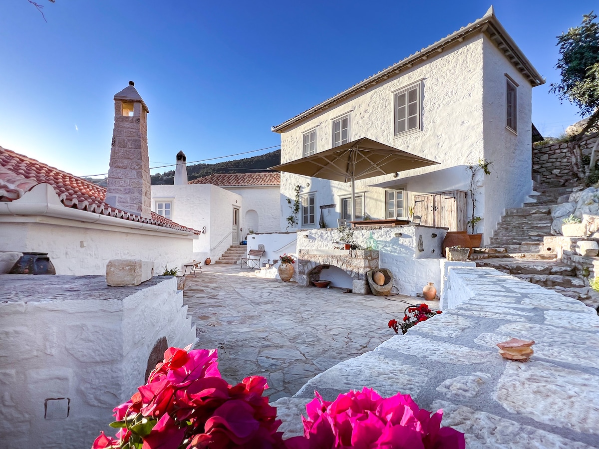 The villa's exterior features a charming white stone façade with a terracotta roof, surrounded by landscaped gardens. A spacious terrace includes a shaded seating area under a large canopy, with potted plants and vibrant flowers enhancing the serene atmosphere.