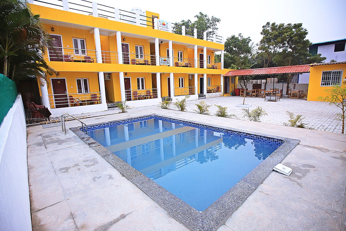 A bright yellow hotel features multiple rooms with balconies overlooking a clean blue pool. Surrounding the pool area, a paved courtyard includes palm plants and outdoor seating arrangements under a shaded canopy. The environment is open and inviting, perfect for relaxation.