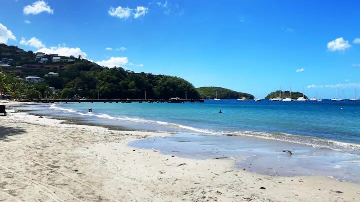 A sandy beach extends alongside calm blue waters, with gentle waves lapping at the shore. In the distance, boats are anchored, and green hills provide a backdrop. The sky is clear with a few clouds, creating a serene coastal atmosphere.