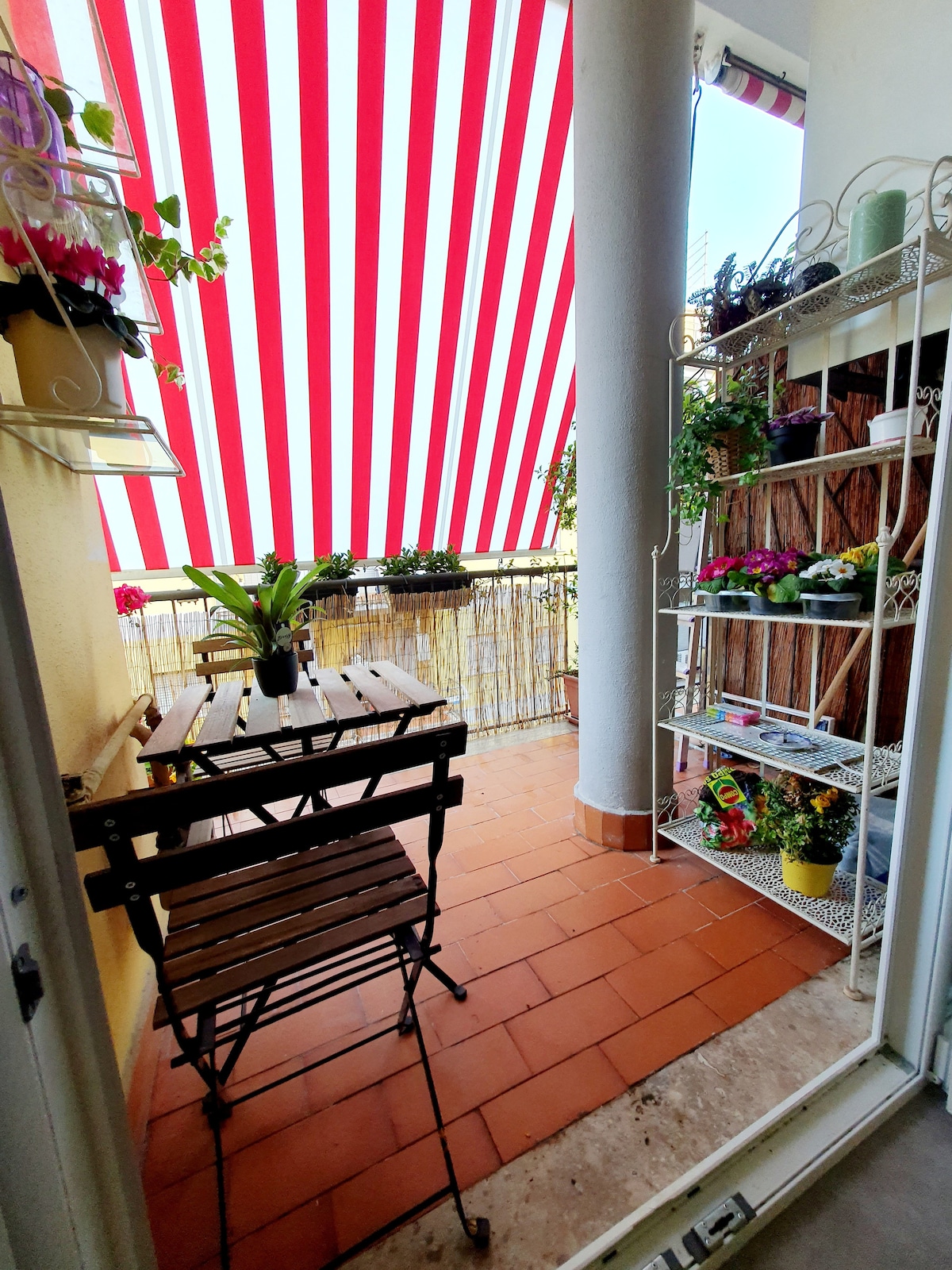 A welcoming terrace is visible, featuring a wooden table and two folding chairs. A sunshade with red and white stripes provides shade. Potted plants adorn the railing and a shelving unit stands against the wall, enhancing the outdoor living space.