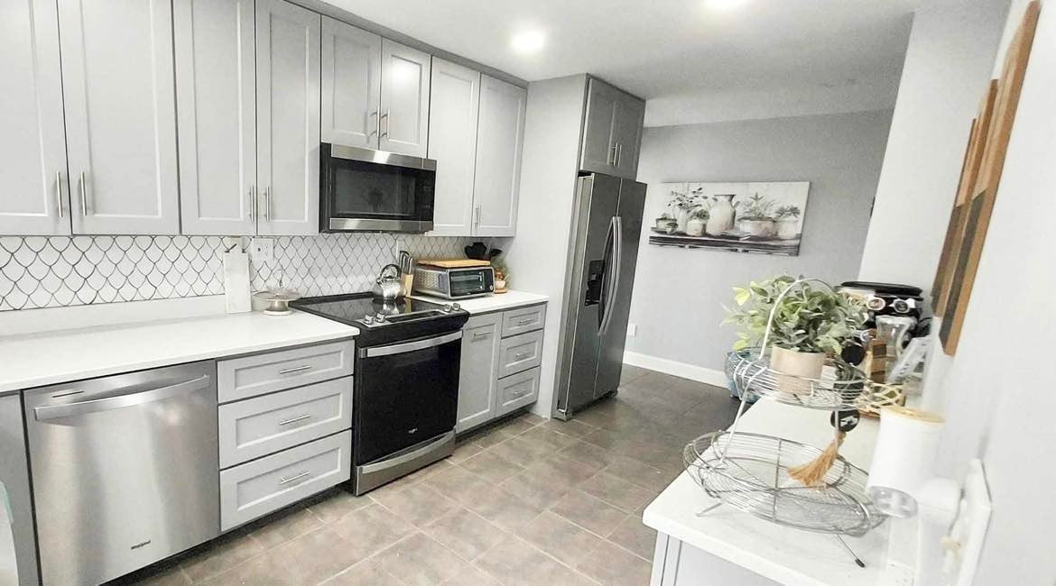 The modern kitchen features sleek gray cabinetry and stainless steel appliances, including a refrigerator and microwave. A white countertop provides ample workspace, complemented by a mosaic tile backsplash. A small table with decorative plants adds a touch of style to the inviting space.
