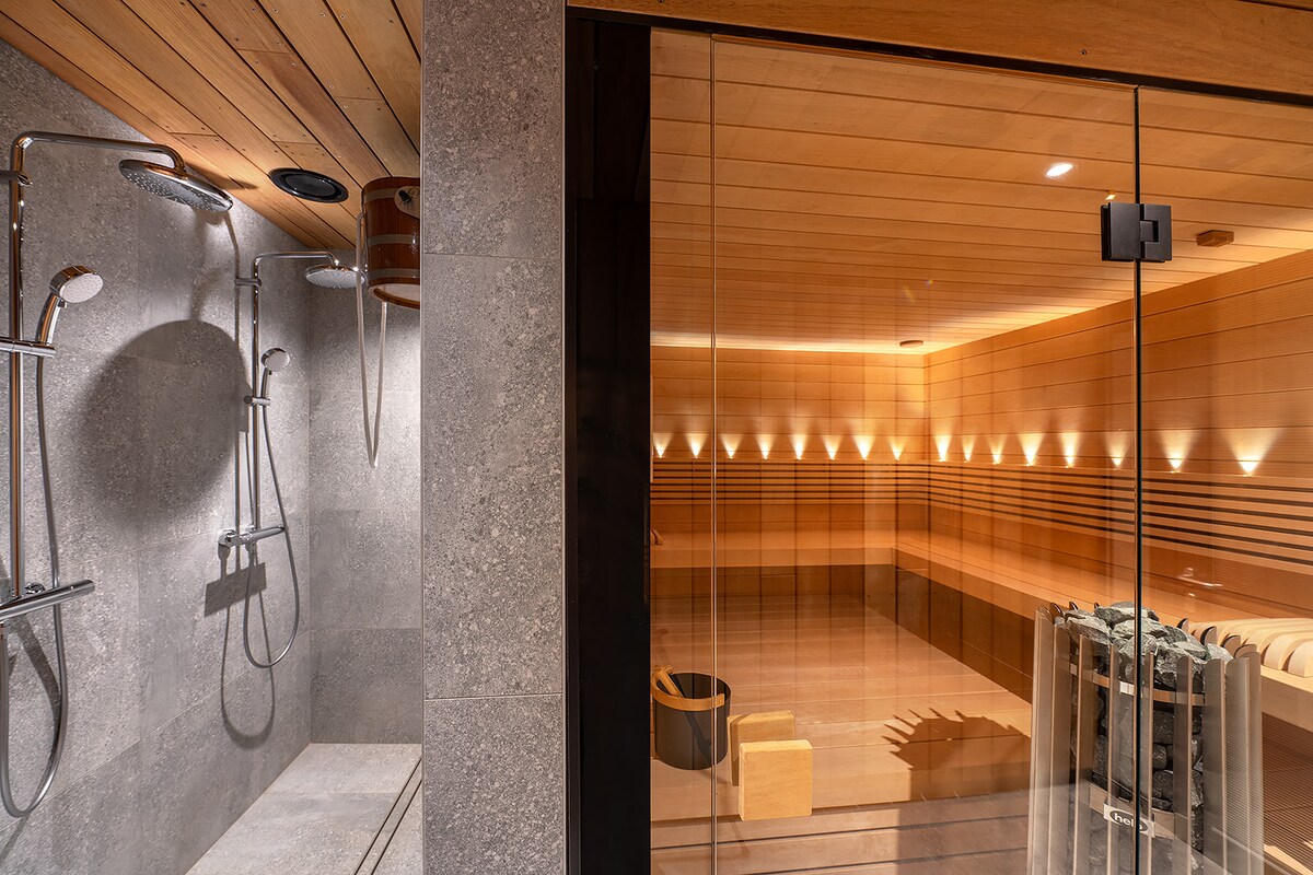 The image showcases a private sauna area featuring wooden walls and ambient lighting. Showers with sleek chrome fixtures are visible on one side, while a spacious relaxation area with minimalist seating can be seen through a glass partition, creating a soothing atmosphere.