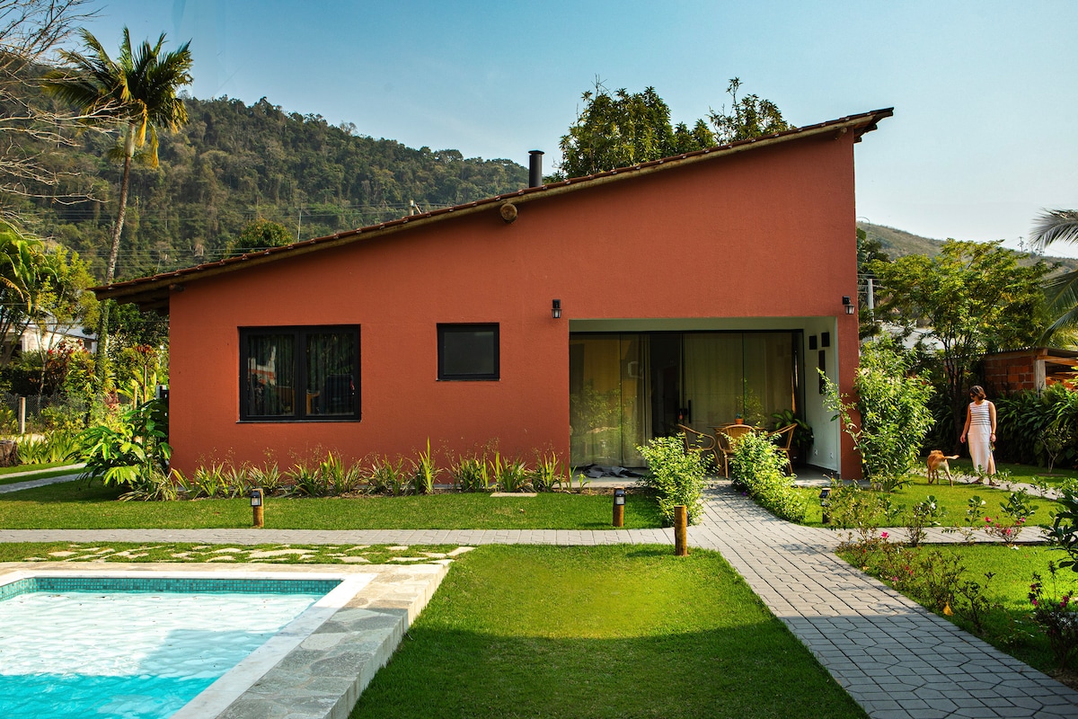 A charming house is shown with a red exterior nestled among lush greenery. The image captures a clear view of the pool in the foreground, with a paved path leading to the entrance. A serene hillside serves as a backdrop, highlighting the peaceful surroundings.