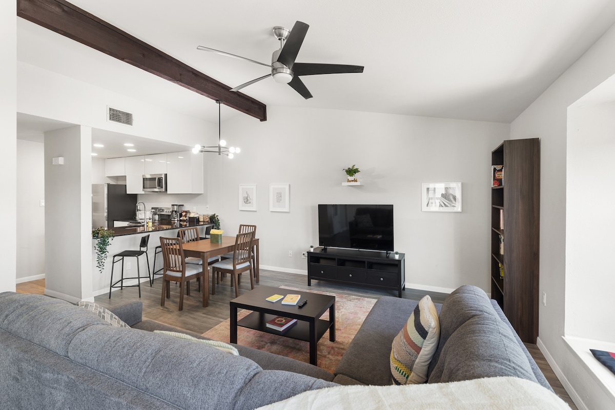 An open living area presents a gray sectional sofa facing a large flat-screen TV. A dining table for six is visible near the modern kitchen, which features dark cabinetry and stainless steel appliances. Natural light enhances the space, with ceiling beams adding architectural detail.