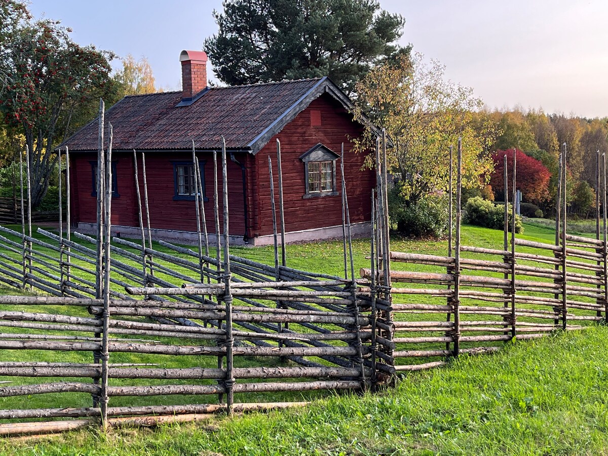 A charming red cottage is framed by a rustic wooden fence, constructed from logs arranged vertically. The surrounding green grass is complemented by trees in vibrant autumn colors and a soft sky, enhancing the natural setting of the property.