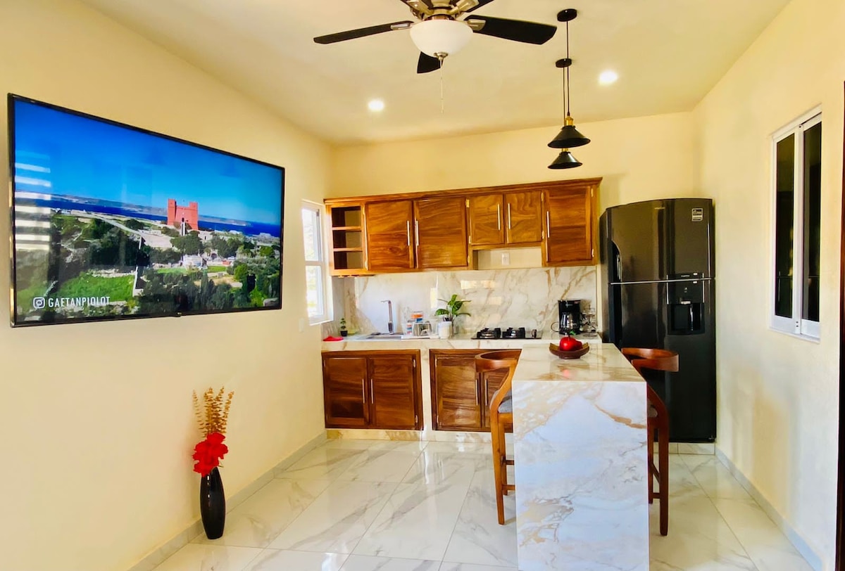A modern kitchen area is highlighted by polished marble flooring and warm wooden cabinetry. A large television mounted on the wall displays scenic views. The kitchen island offers seating for three, with a sleek black refrigerator and a gas stove installed nearby.