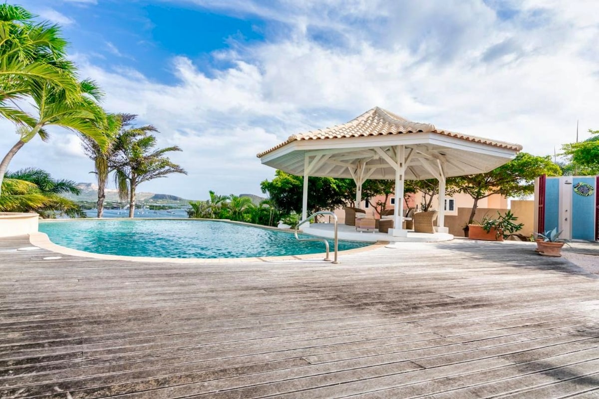 The private pool area is surrounded by lush greenery and palm trees, with a charming gazebo offering shade. The pool's gentle curves reflect the sky, inviting relaxation. Wooden decking encircles the pool, enhancing the open and tropical ambiance.