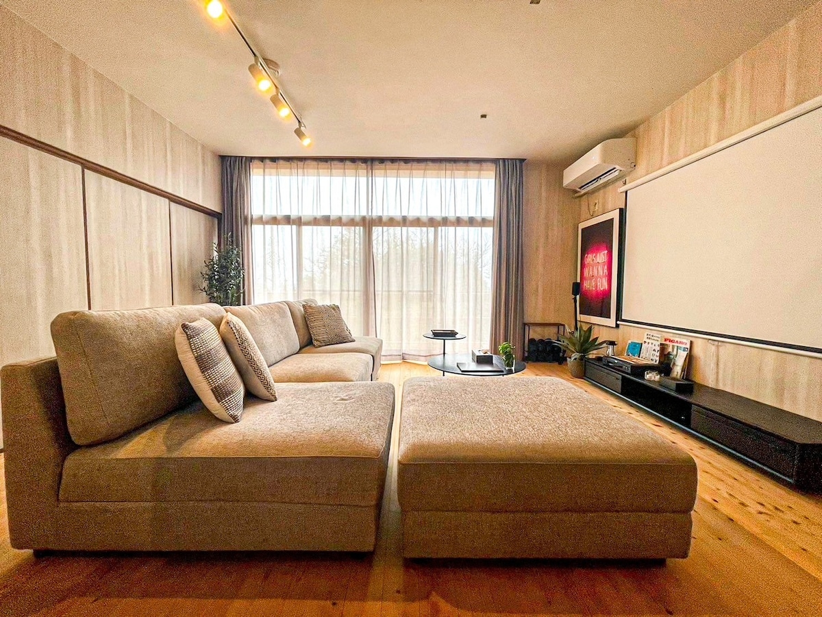 A spacious living area features a large, light-colored sectional sofa and a low coffee table, providing ample seating. Natural light streams in through the extensive windows, with sheer drapes softly filtering the brightness. A projector screen is mounted on the wall for entertainment.