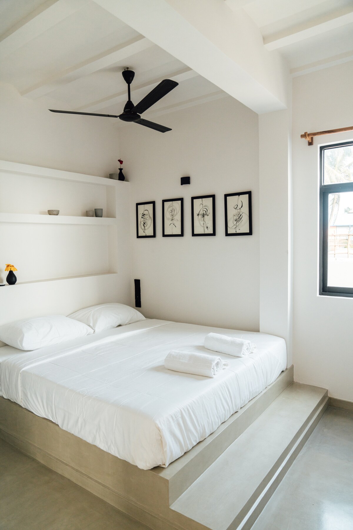 A minimalistic bedroom features a spacious bed with crisp white linens and neatly arranged towels. Artwork is displayed on the wall above the bed, while a fan and natural light enhance the inviting ambiance. A window provides a view of the outside.