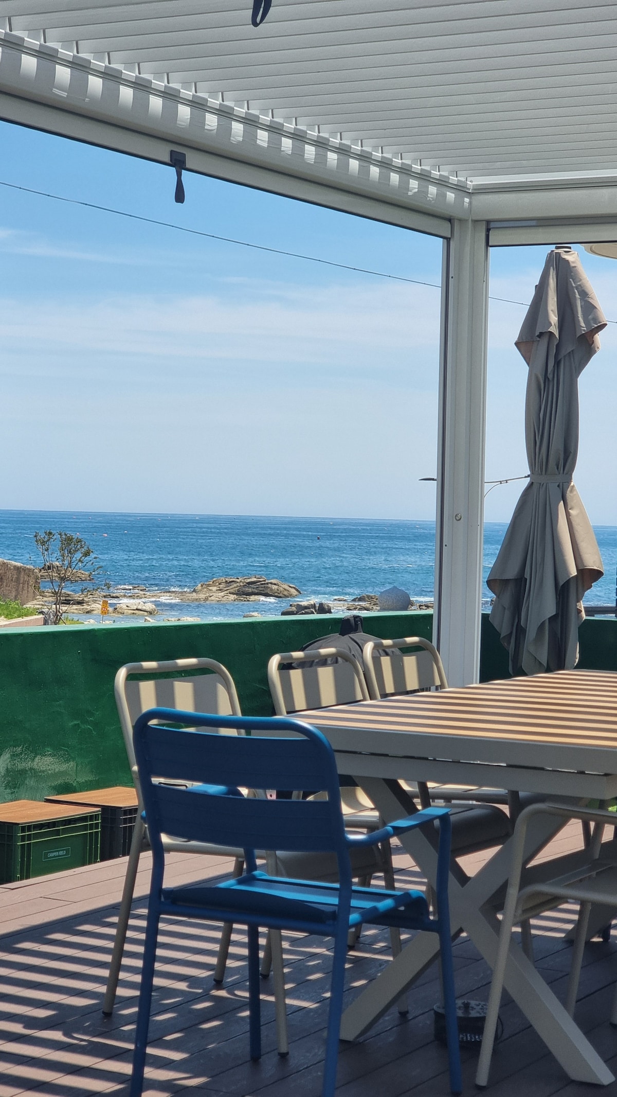 A spacious outdoor dining area is visible under a shaded pergola, featuring a large wooden table surrounded by several chairs. An umbrella stands nearby, with views of the ocean beyond, highlighting the tranquil sea and distant coastline.