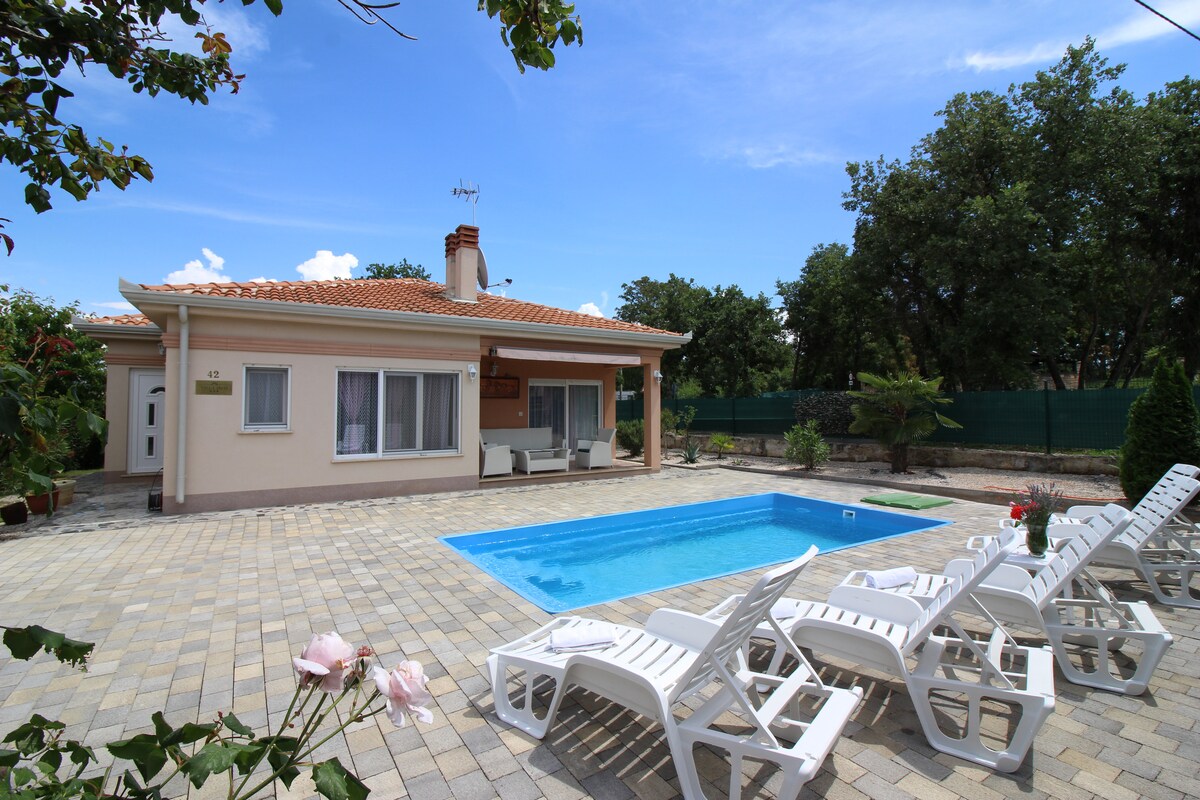 A modern villa is seen with a well-maintained patio surrounding a sparkling pool. Eight lounge chairs are arranged invitingly along the poolside. Large windows are visible on the villa, with a shaded seating area in front, surrounded by greenery and pathways of brick pavers.