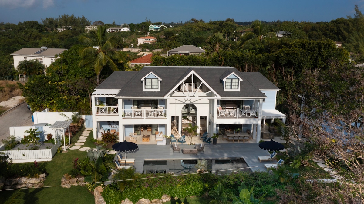 This spacious two-story home features a welcoming exterior with a large, central arched doorway. Balconies adorned with white railings provide an inviting view over the lush tropical landscape, while the tranquil pool area is surrounded by comfortable lounge chairs and vibrant greenery.