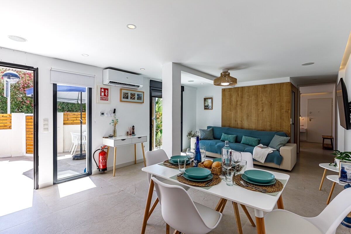 A well-lit living area is showcased, featuring a comfortable sofa and a dining table set for four. Large glass doors open to a private outdoor space, enhancing the connection with nature. Neutral tones and simple decor create a clean, modern aesthetic throughout the room.