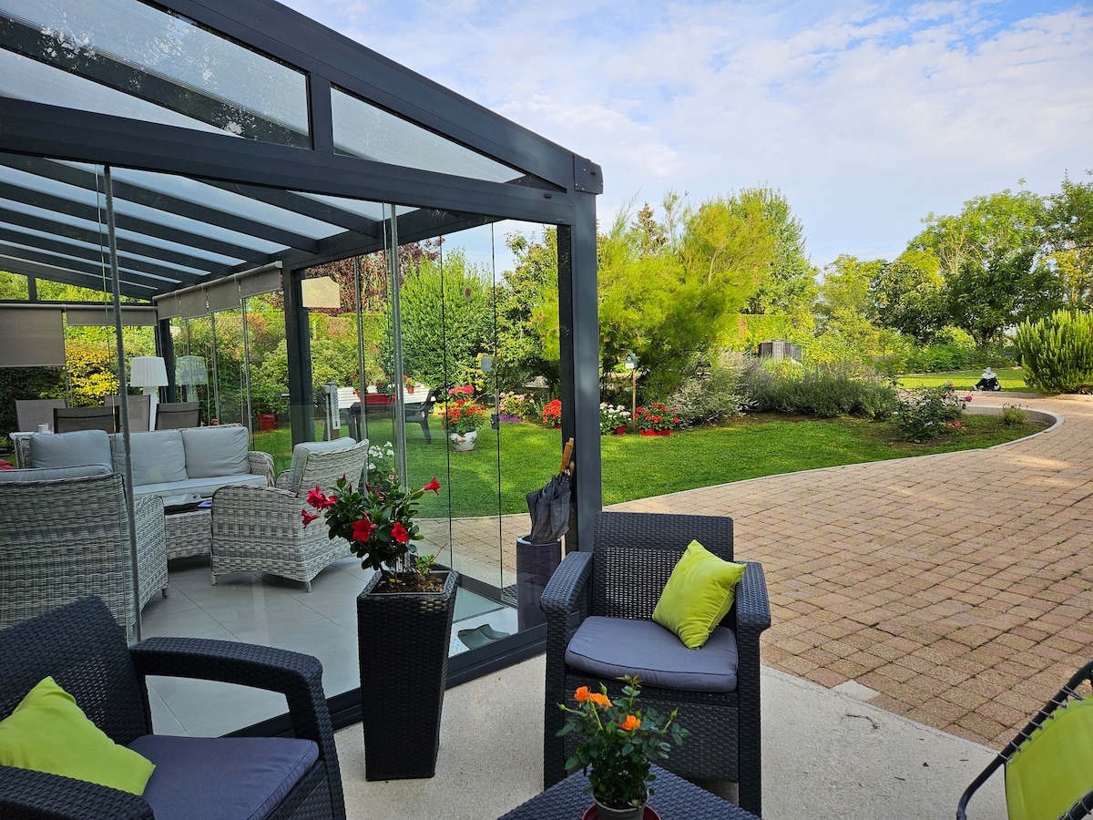 A glass-enclosed terrace features comfortable seating with vibrant cushions. Potted flowers add splashes of color, while a landscaped garden with greenery and blooming plants is visible in the background. A paved pathway meanders through the garden, inviting relaxation and outdoor enjoyment.