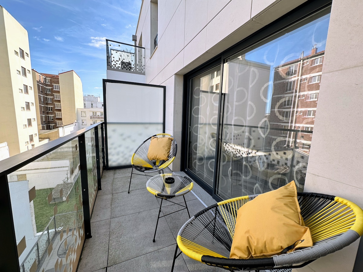 A modern balcony is designed with two stylish chairs featuring yellow cushions. A round table sits in between, providing a space to enjoy refreshments. The glass railing and large sliding doors allow for natural light, creating a bright atmosphere.