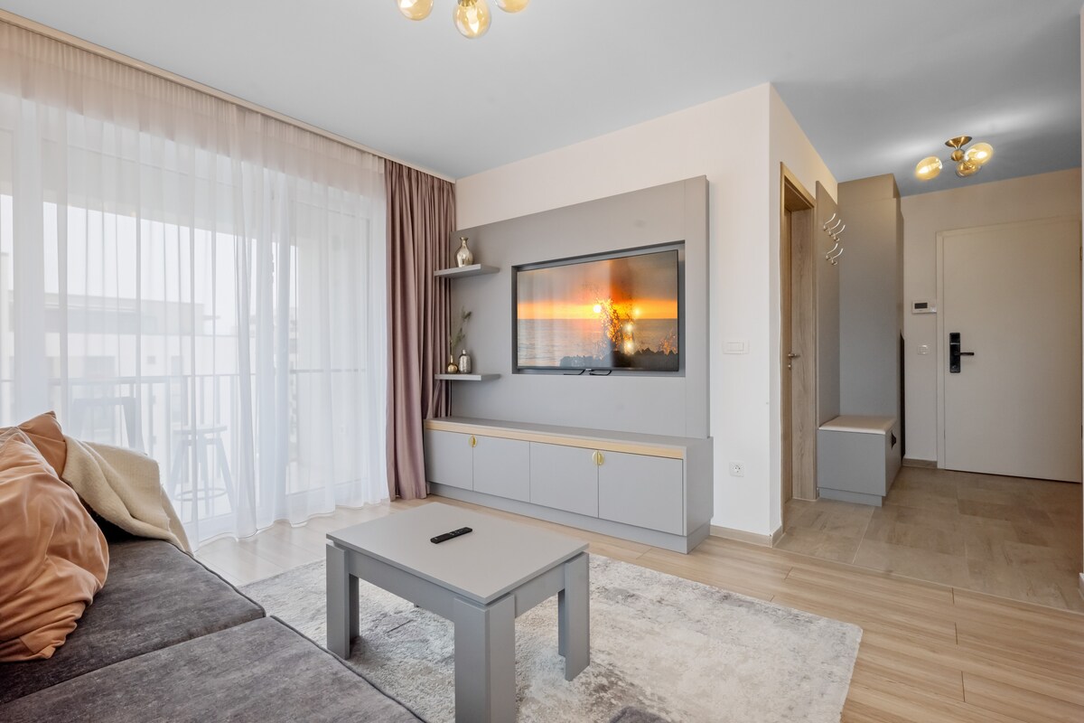 A bright living area features a spacious gray sofa positioned near a light-colored coffee table. A large television is mounted on the wall, showcasing a sunset image. Sheer curtains allow natural light to filter through, creating a relaxed atmosphere.