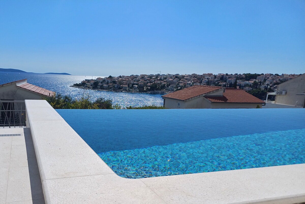 An infinity pool overlooks the sparkling blue waters of Mavarstica Bay, with a scenic view of nearby coastal towns in the distance. The pool area is lined with a sleek, light-colored stone terrace, creating a serene space for relaxation.