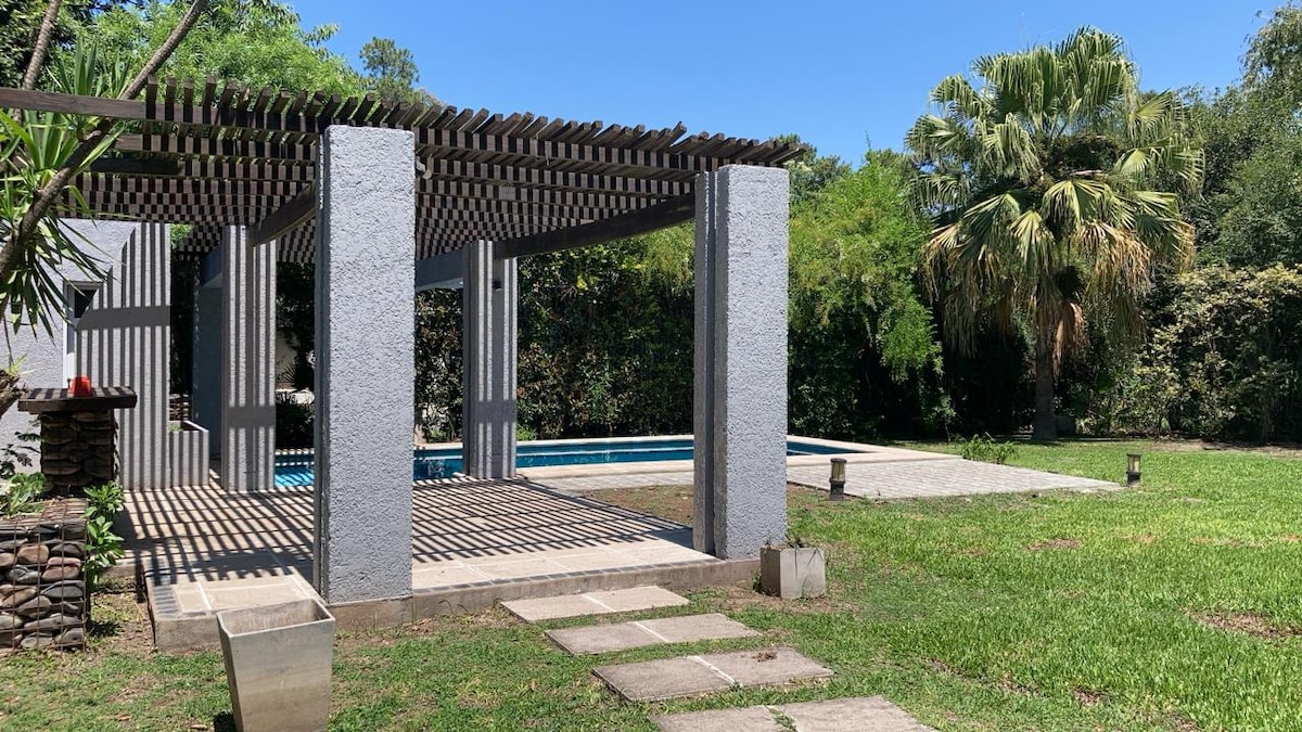 An outdoor patio features a pergola with striped wooden slats, providing shade over a stone-tiled area. Lush greenery surrounds the space, including tall palm trees and neatly trimmed grass. A clear blue pool is visible in the background, offering a refreshing focal point.