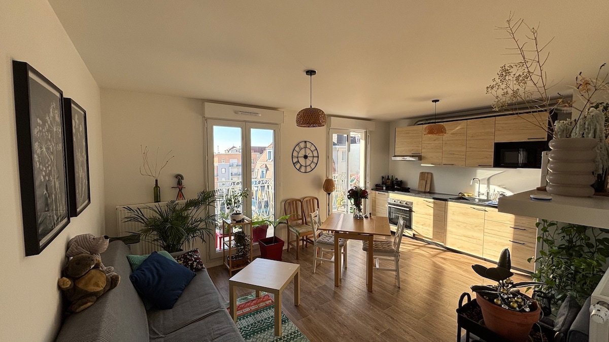 An open living area is showcased with a spacious sofa and a wooden dining table. Natural light fills the space through large windows, highlighting various green plants. Modern kitchen appliances are visible alongside warm wooden cabinetry, creating a cozy and functional environment.