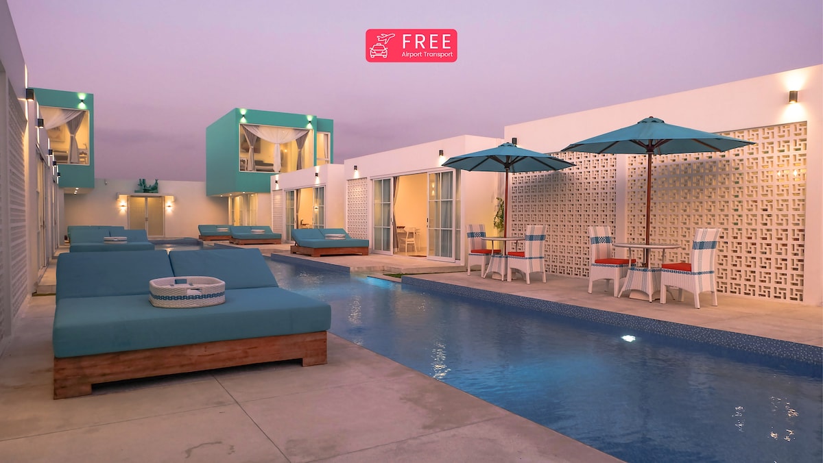 A serene pool area is showcased, featuring a long pool bordered by comfortable lounge beds. Umbrellas offer shade over dining tables, while contemporary villa units with glass doors provide an open and airy environment. Soft evening light bathes the scene in a tranquil setting.