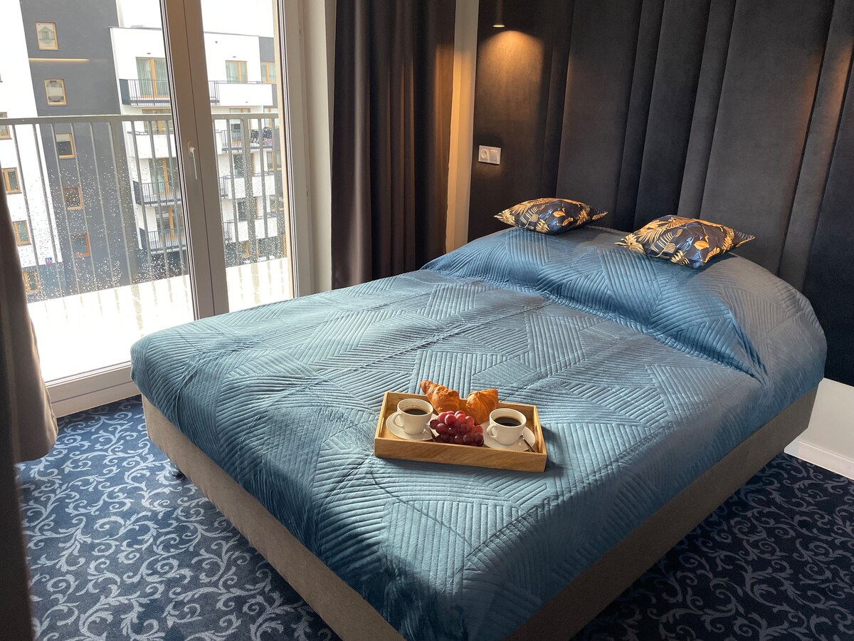 A cozy sleeping area features a spacious bed with a textured blue coverlet. Two pillows with a floral design sit atop the bed. A wooden tray displays two cups and a small assortment of pastries. Natural light enters through sliding glass doors leading to a balcony.