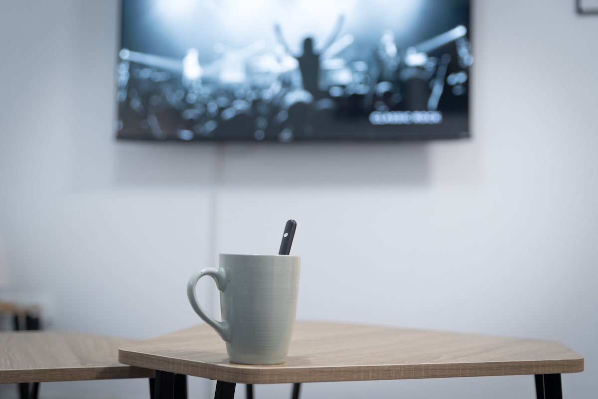 A wooden table is featured in the foreground with a light-colored cup and spoon placed on it. Behind, a large flat-screen TV displays a monochromatic image of an audience enjoying a performance, showcasing an inviting space for relaxation.