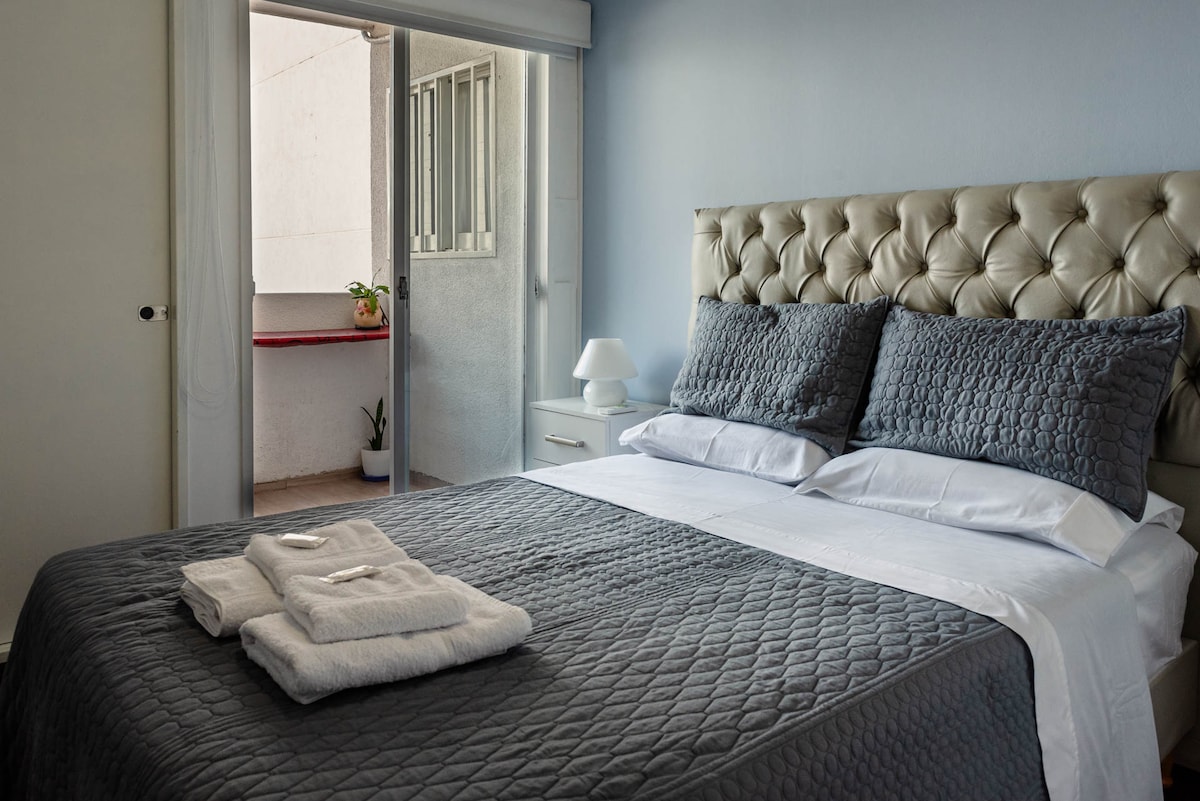A cozy bedroom features a spacious bed with a tufted headboard, layered with a quilted gray blanket. Two decorative pillows sit at the head of the bed, and a small lamp is positioned on a nightstand. Natural light enters through glass doors leading to a small balcony.