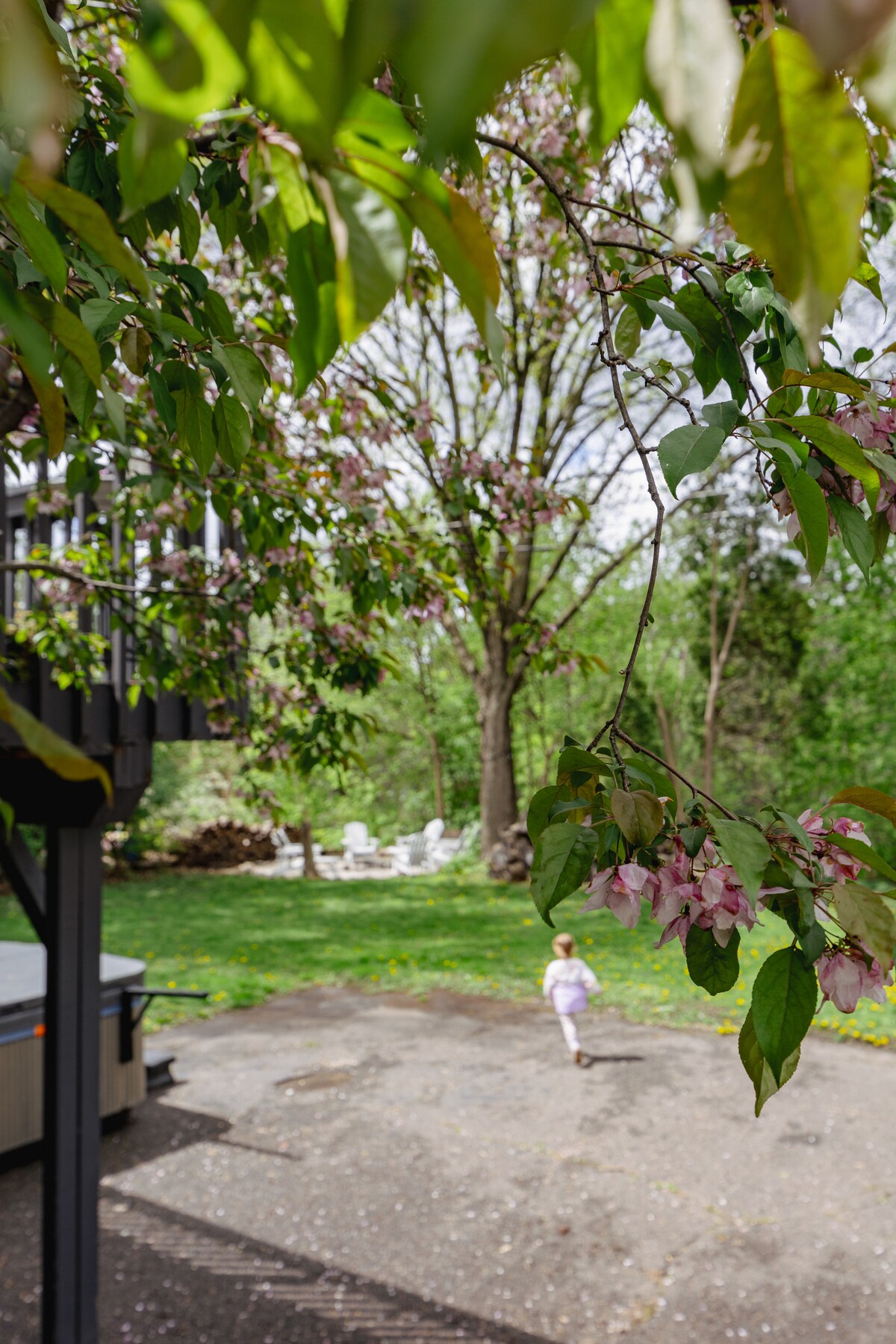 A child runs along a paved area surrounded by lush greenery and blooming trees. In the background, a hot tub is visible, along with a small seating area of white chairs nestled in the yard. Delicate pink flowers adorn the branches partially framing the view.