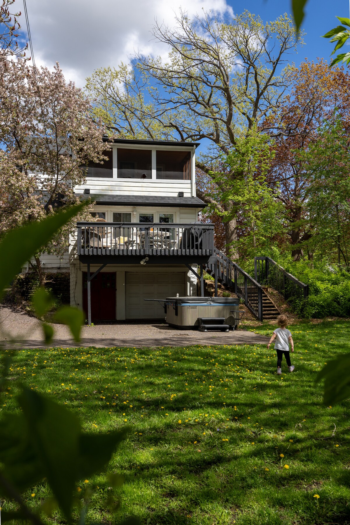The cottage is framed by lush greenery and blooming trees, showcasing a spacious deck with black railings and stairs leading up to a welcoming entrance. A hot tub is positioned beside the house, while a child can be seen playing on the grassy lawn.