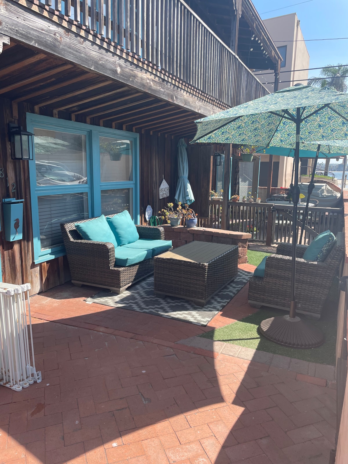 An inviting outdoor patio features a wicker seating arrangement with turquoise cushions and a coffee table. A patterned area rug anchors the space, and a large umbrella provides shade. Lush greenery and decorative elements enhance the tranquil atmosphere.