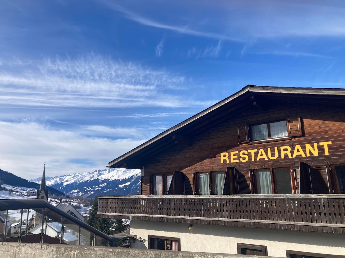 A wooden building features a prominent yellow sign reading 'RESTAURANT.' The exterior showcases multiple balconies with wooden railings. A backdrop of snow-capped mountains and a clear blue sky enhances the setting, while a portion of the town is visible below.