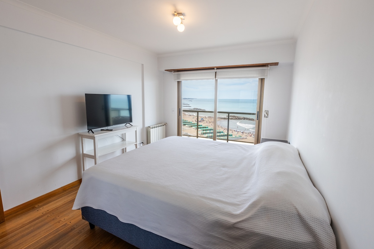 A spacious sleeping area is presented, featuring a large bed with a smooth white coverlet. A television is positioned on a white stand, while large windows offer an unobstructed view of the beach and ocean beyond.