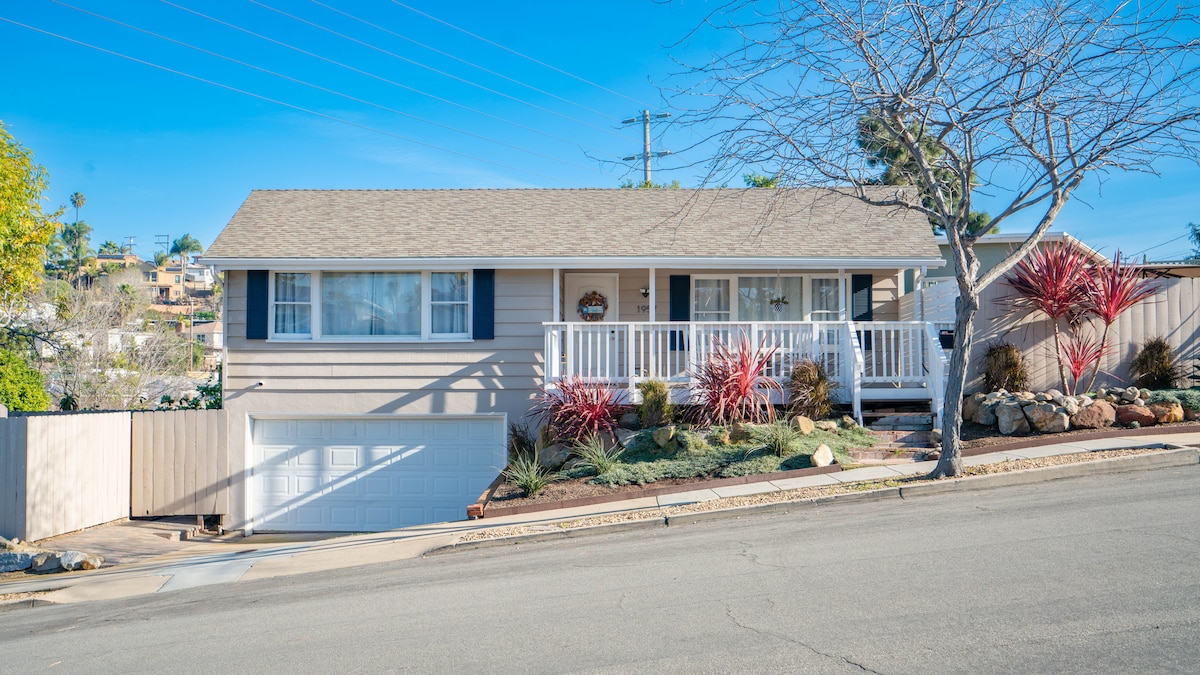 The exterior of the vacation home features a light-colored facade with a welcoming front porch adorned with white railings. A well-maintained lawn and colorful plants enhance the curb appeal. The driveway leads to a garage, and the surrounding neighborhood is visible on a gentle slope.