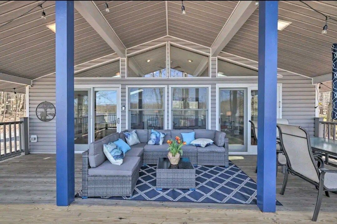 A partially covered outdoor deck features comfortable seating arranged around a central table, adorned with a floral centerpiece. Large windows offer views of the lake, while a ceiling fan provides a cool breeze in the open space.