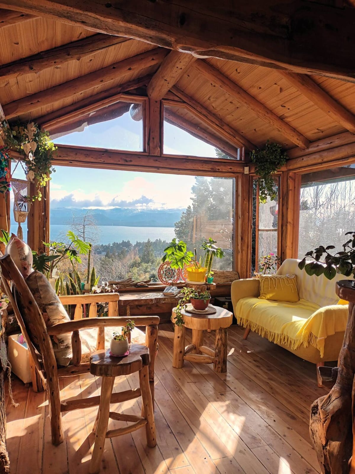 A sunlit sitting area features large windows that provide a view of the lake and surrounding nature. A yellow draped sofa complements wooden furniture, including a small table and rustic chairs. Various plants are placed on the table and windowsills, adding a touch of greenery.