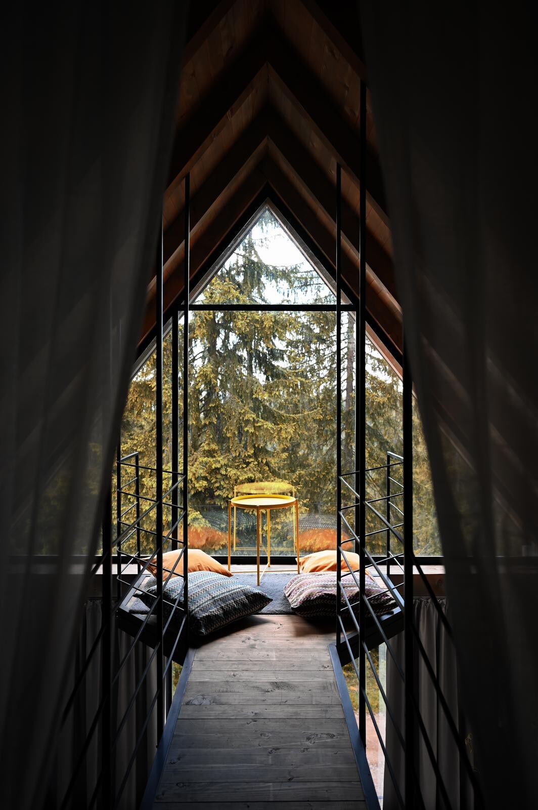 A distinctive architectural design is featured, showcasing an angled ceiling and large windows that offer a view of the surrounding trees. A narrow walkway leads to a small table adorned with cushions, creating a relaxed spot to enjoy nature.