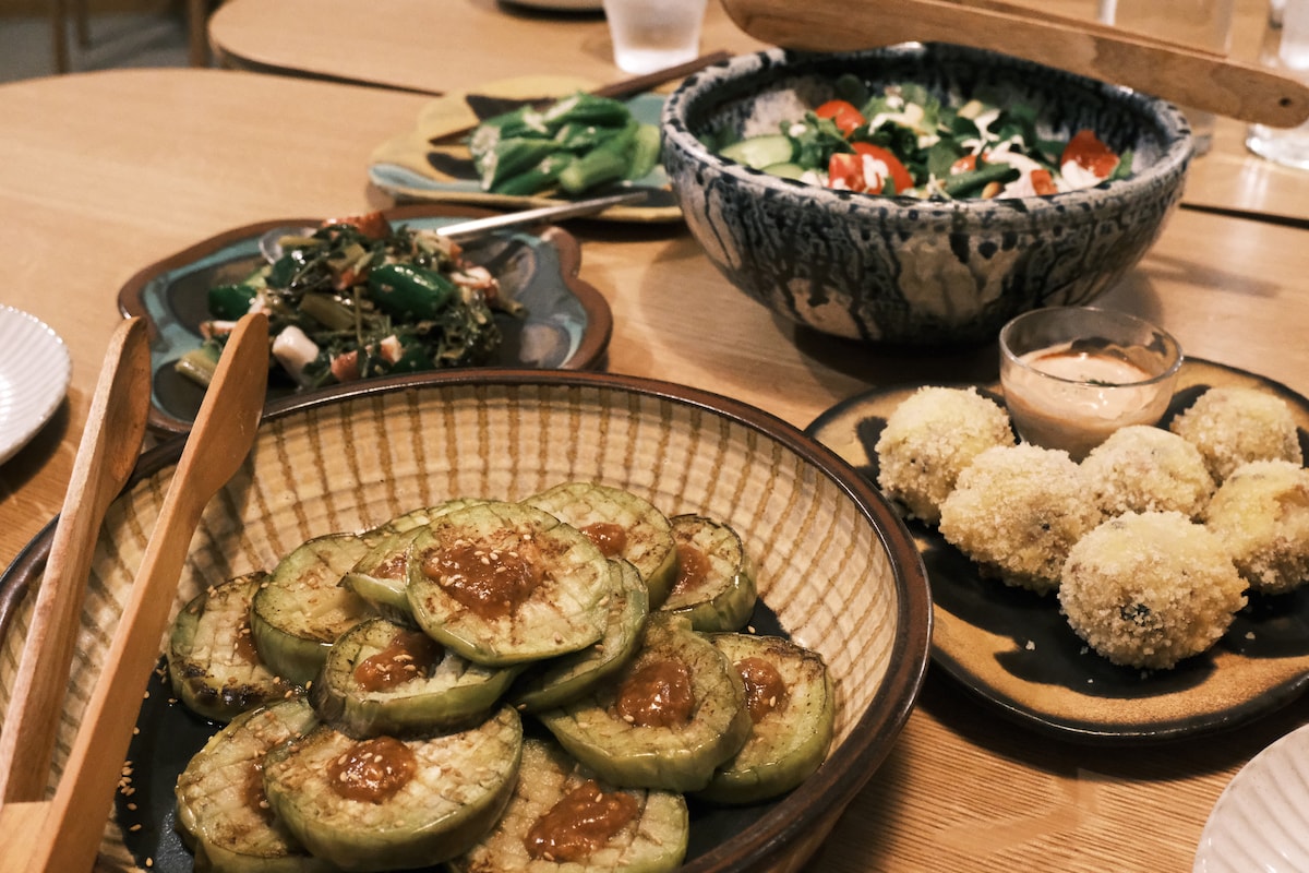 An assortment of dishes is displayed on a wooden table. Sliced eggplant with sauce occupies the foreground, while golden-brown breaded balls are presented in another dish. A colorful salad featuring greens and tomatoes is visible alongside a platter of stir-fried vegetables, highlighting a diverse meal selection.