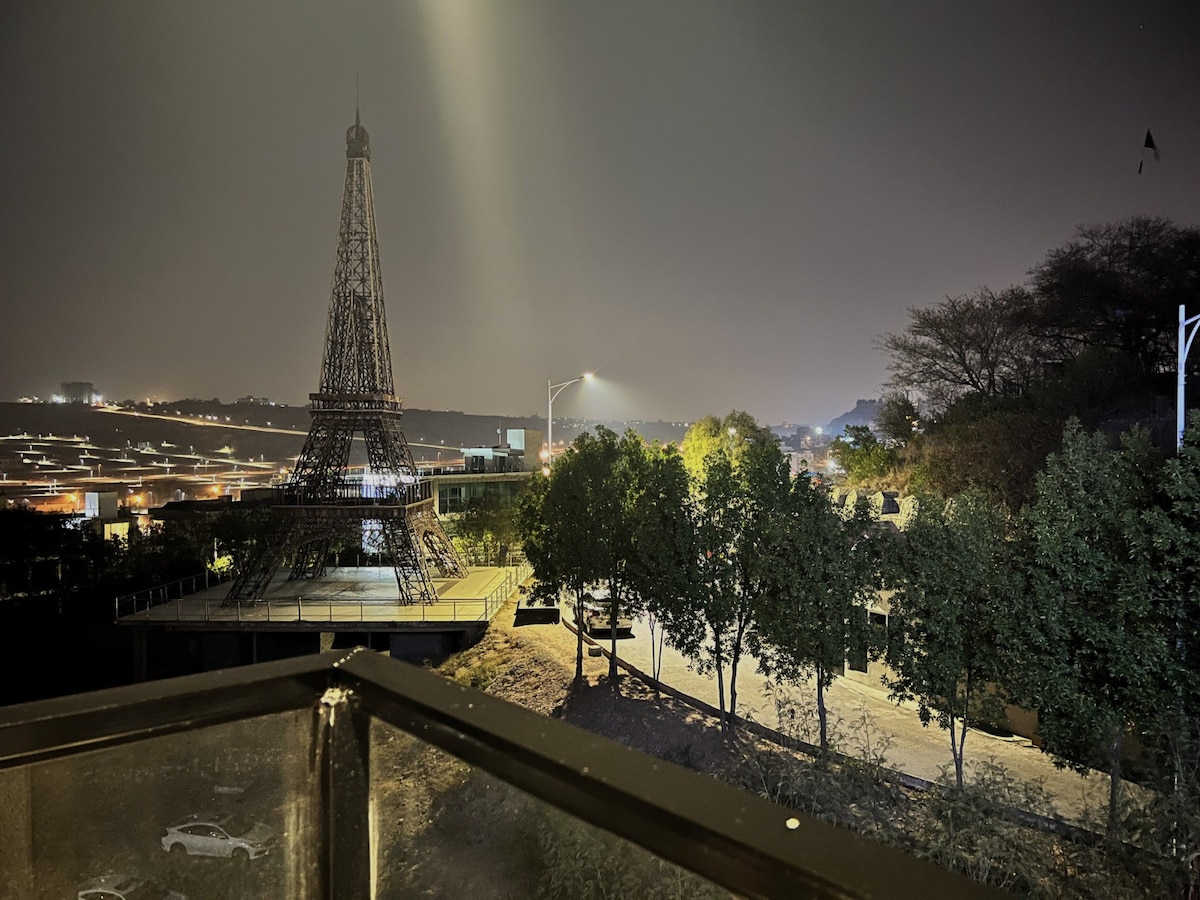 A nighttime view features a replica of the Eiffel Tower, illuminated against the dark sky. Surrounding greenery provides a contrast to the structure, while dim lighting enhances the serene atmosphere. The scene offers a glimpse of the nearby landscape, subtly highlighted by ambient light.