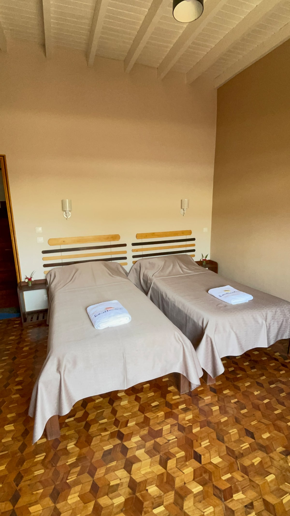 A well-lit bedroom features two neatly made twin beds, each covered with light-colored bedding. The room showcases a wooden floor with a distinctive pattern, and minimalist wall sconces provide subtle illumination.