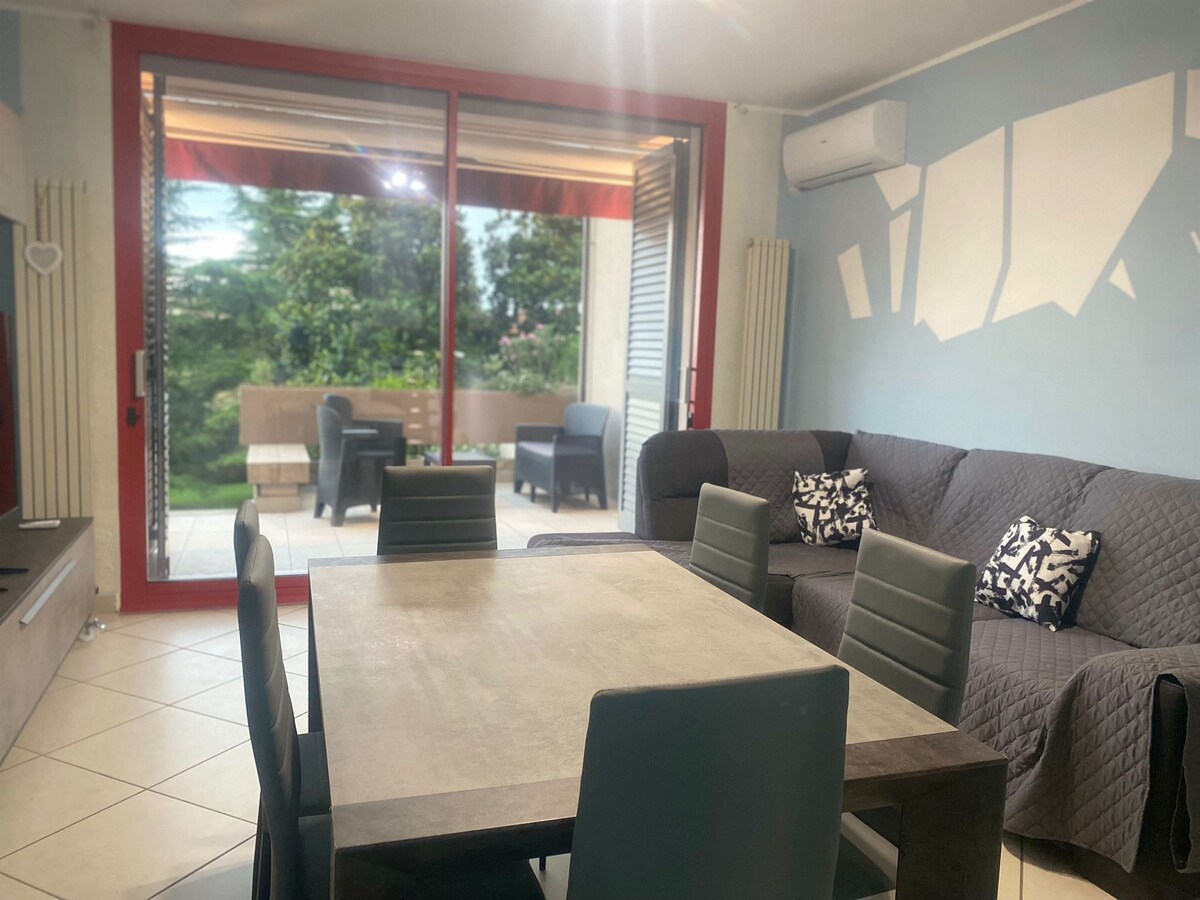 A spacious living area features a modern dining table surrounded by six chairs. A comfortable sofa is positioned against the wall, and large glass doors open to a furnished terrace, offering views of the greenery outside.