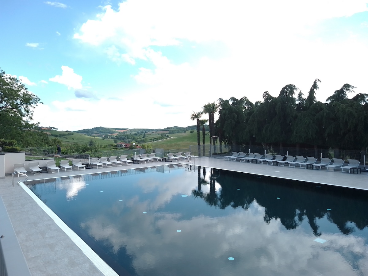 A large, heated swimming pool glistens under a partly cloudy sky, reflecting the surrounding greens of the hills. Sun loungers are arranged neatly around the pool area, providing ample space for relaxation. Towering trees complement the peaceful landscape in the background.