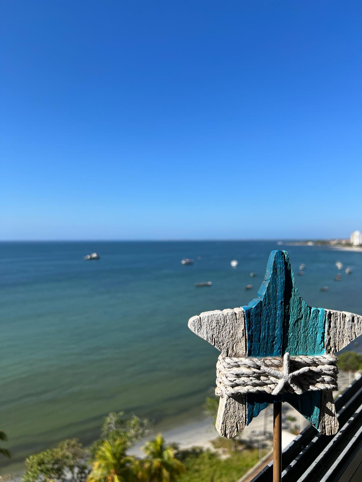 A decorative star shaped object is positioned in the foreground, with a textured blue and white finish. The calm sea is seen in the background, reflecting a clear blue sky, while boats can be spotted gently bobbing on the water.