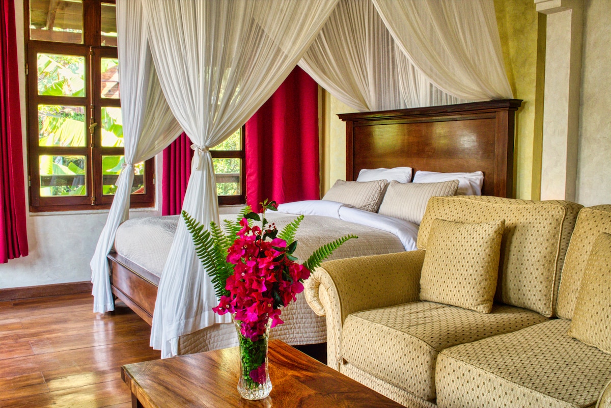 A tranquil bedroom features a queen-size bed adorned with white linens, draped with light, flowing curtains. A cozy sofa and a wooden coffee table enhance the inviting atmosphere. Bright red curtains frame the windows, allowing natural light to illuminate the space while a vibrant floral arrangement adds a pop of color.